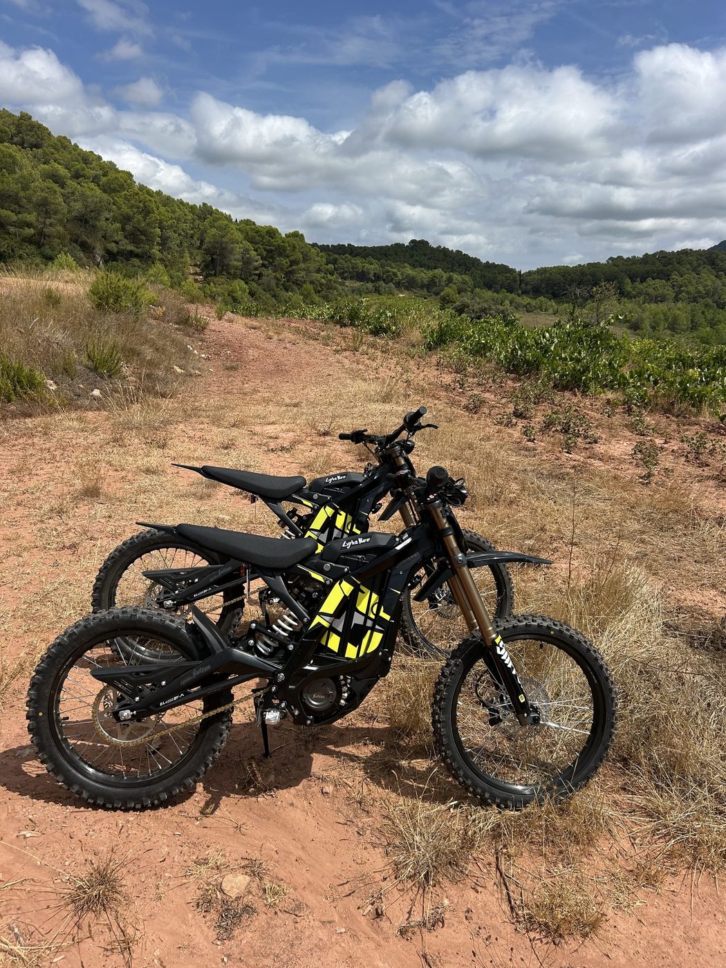 Two electric dirt bikes on an open red track with wooded hills behind